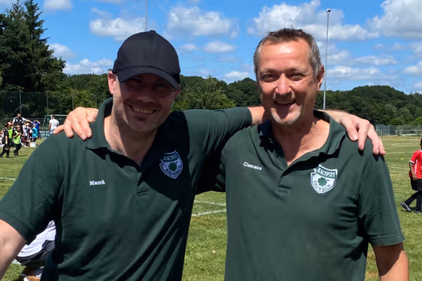 Zwei Männer in grünen Polohemden stehen eng beieinander auf einem Sportfeld, lächelnd und posierend. Im Hintergrund ist ein blauer Himmel zu sehen, gespickt mit wenigen Wolken, sowie grüne Wiesen und Sportaktivitäten.