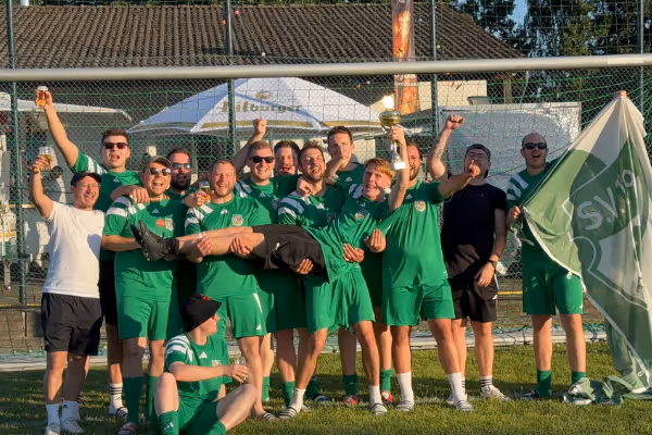 Eine Gruppe von Männern und Frauen in grünen Sporttrikots posiert fröhlich auf einem Fußballfeld. Einige halten eine Flagge, während andere ihre Arme in die Luft heben, um ihren Sieg zu feiern. Im Hintergrund sind Sporteinrichtungen sichtbar.