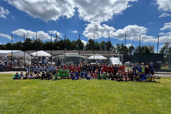 Eine Gruppe von Sportlern in verschiedenen farbigen Trikots versammelt sich auf einem Grasfeld unter blauem Himmel mit weißen Wolken. Im Hintergrund sind Zuschauer und Sporteinrichtungen zu sehen.