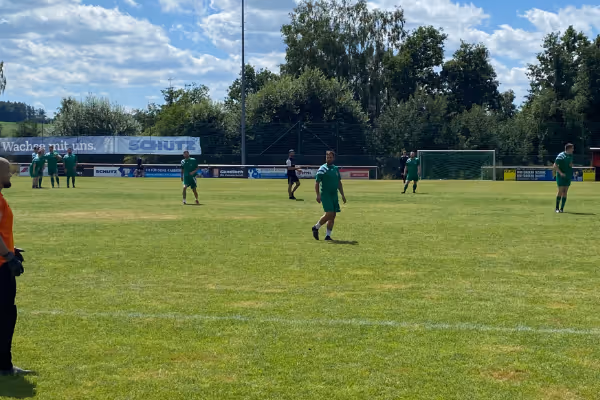 Fußballspiel auf einem sonnigen Platz mit Spielern in grüner Uniform, die aktiv während des Spiels agieren. Im Hintergrund sind Bäume und das Vereinsgebäude sichtbar.
