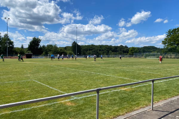 Eine weitläufige Fußballplatzansicht unter einem blauen Himmel mit weißen Wolken, auf dem Spieler aktiv einem Ball nachjagen. Im Hintergrund sind Bäume und Zuschauer am Spielfeldrand sichtbar.
