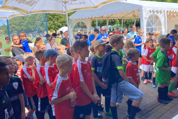 Eine Gruppe von Kindern in roten Trikots steht in einer Reihe auf einem Sportplatz, umgeben von Zuschauern in einem festlichen Ambiente mit Zelten und Bäumen im Hintergrund.