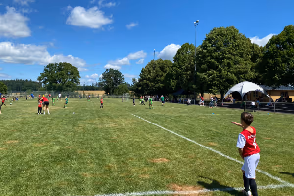 Ein Kind in einem roten Trikot steht am Rand eines Fußballfeldes und beobachtet das Spiel. Im Hintergrund sind Spieler und Zuschauer vor einer blau-himmeligen Landschaft zu sehen, umgeben von grünen Bäumen.
