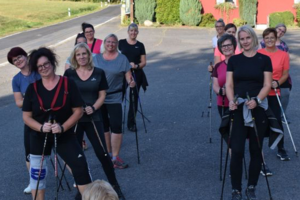 Eine Gruppe von Frauen steht in einer Reihe auf einer Straße im Freien und hält Nordic-Walking-Stöcke in den Händen. Sie tragen sportliche Kleidung und lächeln freundlich in die Kamera. Im Hintergrund sind grüne Bäume und ein rotes Gebäude sichtbar.