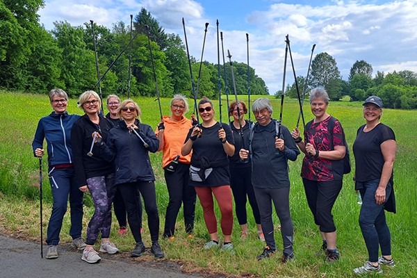 Eine Gruppe von Frauen steht in einer Reihe auf einer Straße im Freien und hält Nordic-Walking-Stöcke in den Händen. Sie tragen sportliche Kleidung und lächeln freundlich in die Kamera. Im Hintergrund sind grüne Bäume und ein rotes Gebäude sichtbar.