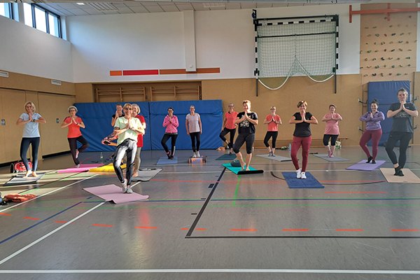 Eine Frau in einer Yoga-Position auf einer pinken Matte in einem hellen Raum. Sie hat ihre Arme weit ausgestreckt und ist entspannt. Der Raum ist minimalistisch, mit einem hellen Boden und einer ruhigen Atmosphäre.