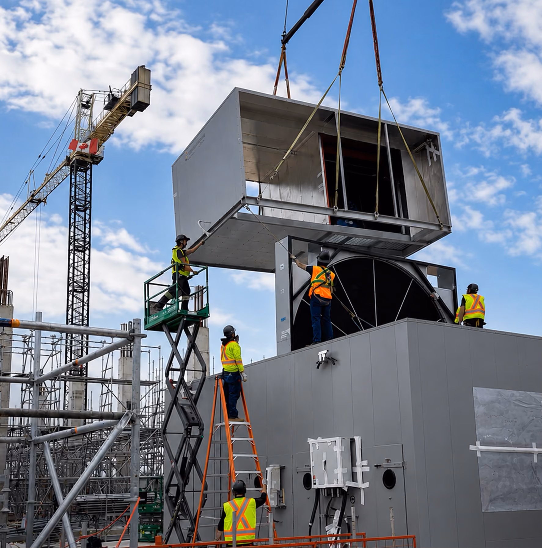 Construction workers with safety gear guide a large metal module being lifted by a crane on a building site.
