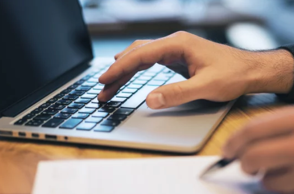 Person typing on a laptop keyboard with a hand holding a pen near a paper on a wooden desk.