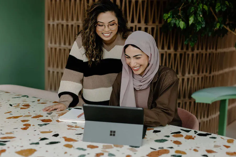 two females working on a laptop, smiling set in a modern office.