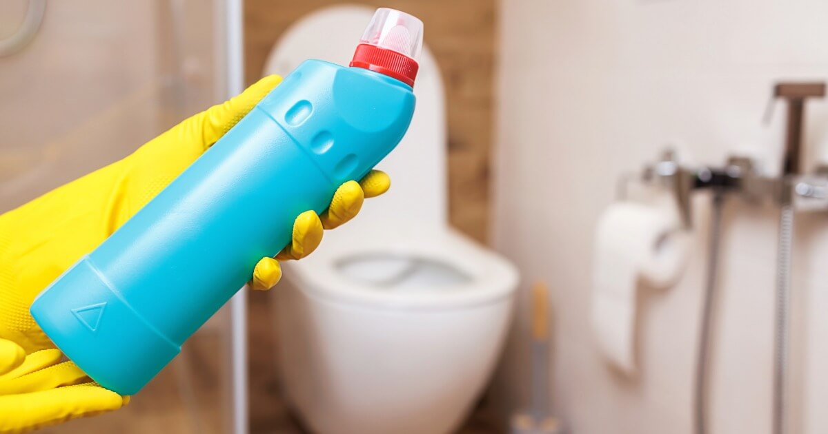 Gloved hand holding toilet cleaner in bathroom, with toilet and paper roll in the background.