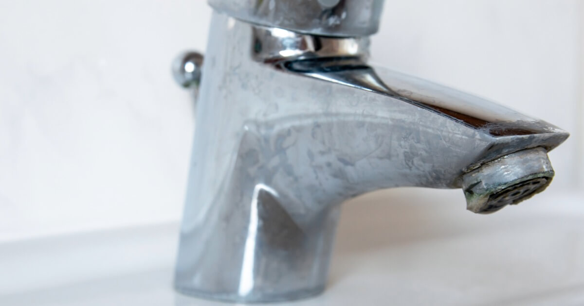 Close-up of a chrome bathroom faucet with visible hard water stains, set against a white tiled background.