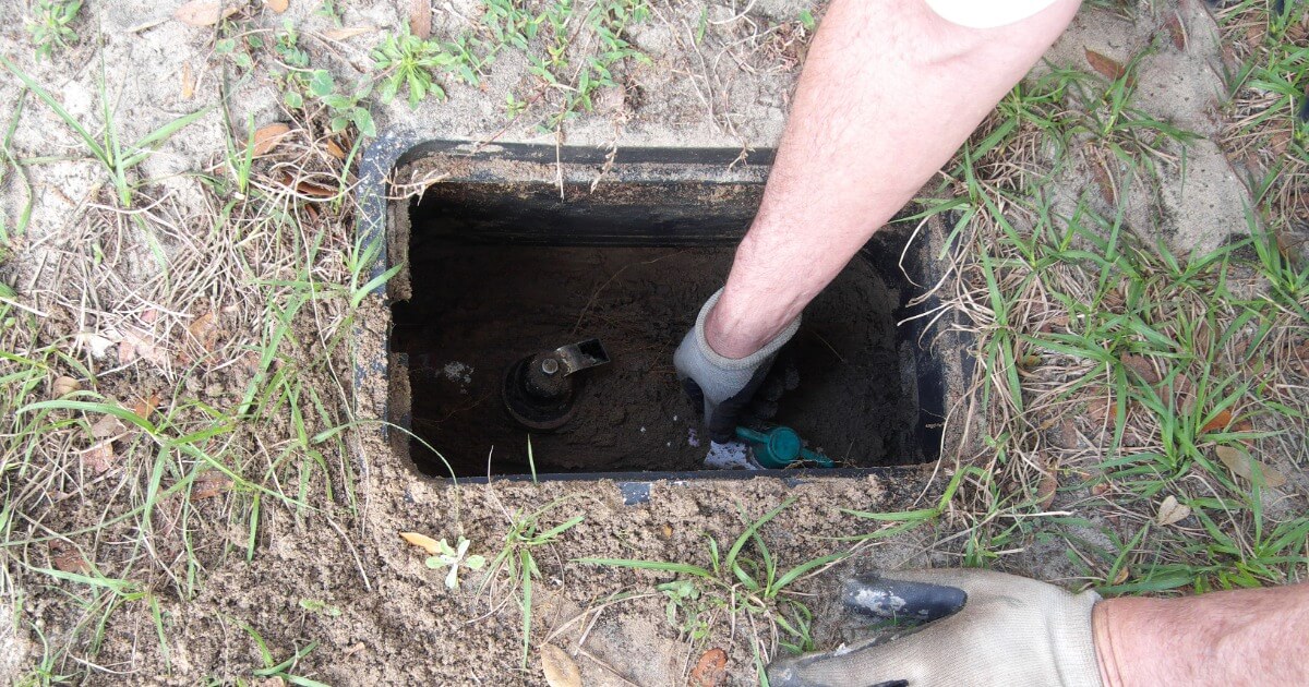 Person checking emergency shutoff valve in a grassy area with a gloved hand inside the meter box.