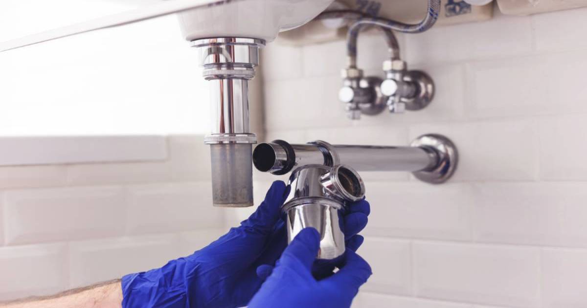 Plumber wearing blue gloves inspects piping under bathroom sink. 