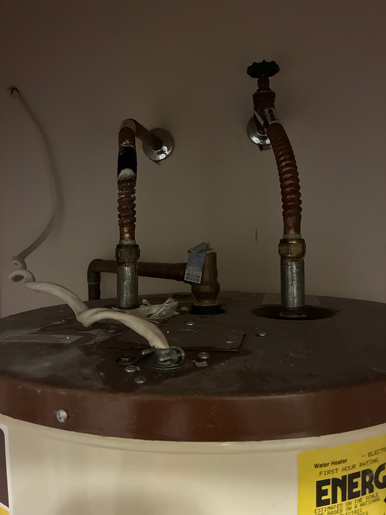 Top view of a water heater with rusted metal pipes and two corrugated copper tubes connected to the top panel against a plain wall.