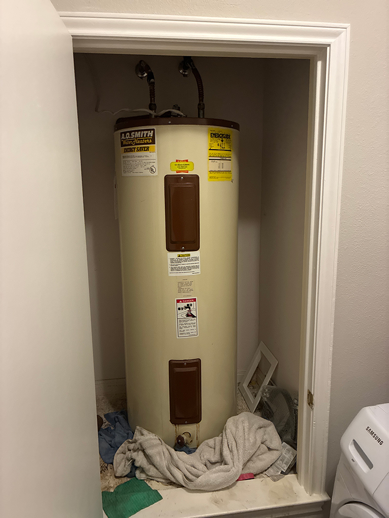 A cylindrical A.O. Smith water heater with brown panels in a small closet surrounded by towels and rags on the floor.