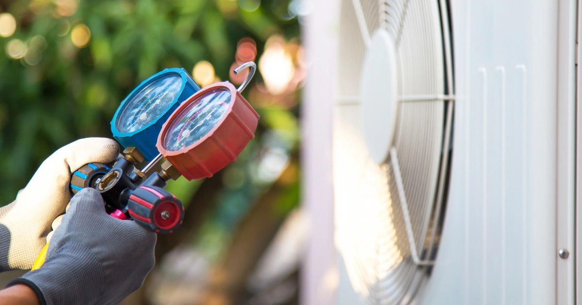 Gloved hands holding blue and red AC pressure manifold gauges in front of an outdoor air conditioner unit.