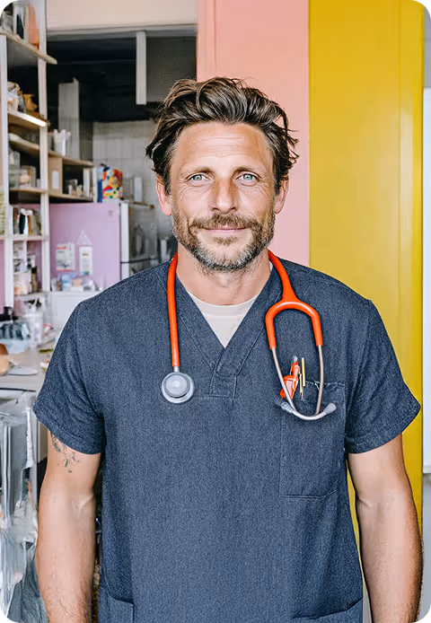 Un homme souriant en tenue médicale bleue avec un stéthoscope orange autour du cou, debout dans un intérieur coloré.