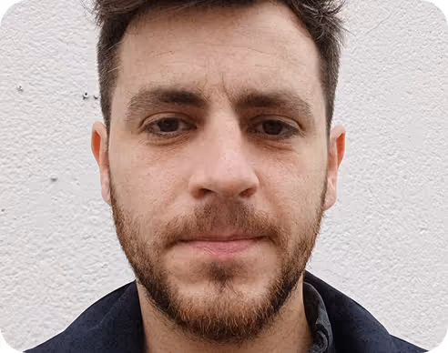 Portrait d'un homme avec barbe et cheveux courts, portant un manteau foncé devant un mur blanc.