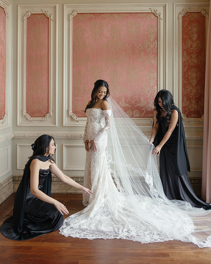 Bride in an off-shoulder lace wedding gown with a long veil being assisted by two bridesmaids in black dresses in an ornate room with red and gold wallpaper.