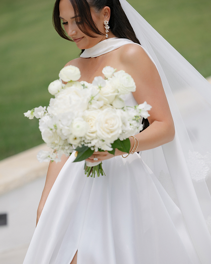 Bride in a white gown holding a bouquet of white flowers, wearing a veil and pearl earrings, looking down with a gentle smile.