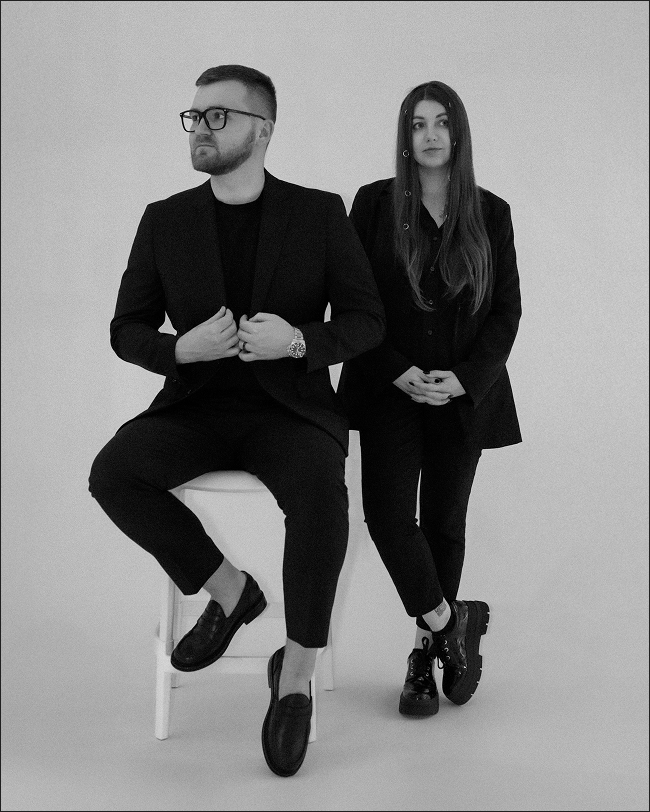 Black-and-white photo of a man sitting on a stool and a woman standing beside him, both dressed in black attire against a plain background.