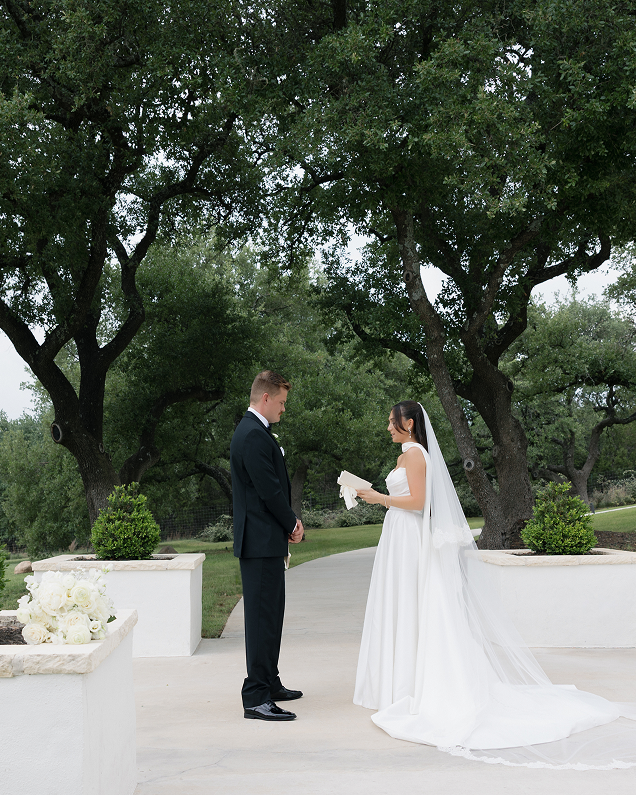 Bride in white dress and veil reading vows to groom in black suit outdoors under large trees.