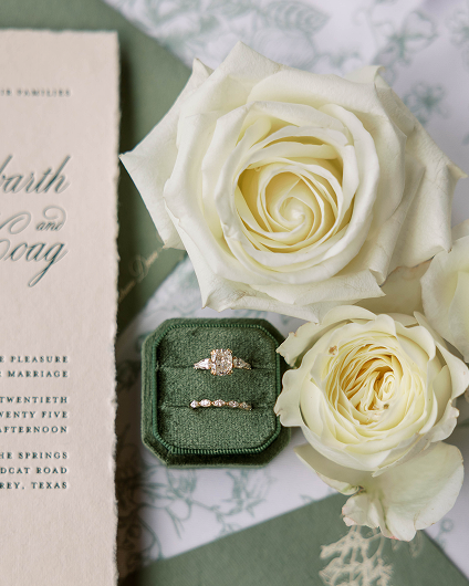 Green velvet ring box with two diamond rings next to two white roses and a wedding invitation.