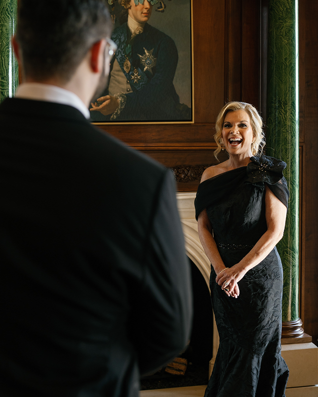 Smiling woman in an elegant black dress standing indoors, facing a man in a suit with a framed portrait hanging on the wood-paneled wall behind her.