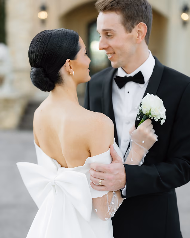 Bride with a large bow on her white dress embracing groom in black tuxedo holding a white rose boutonniere.