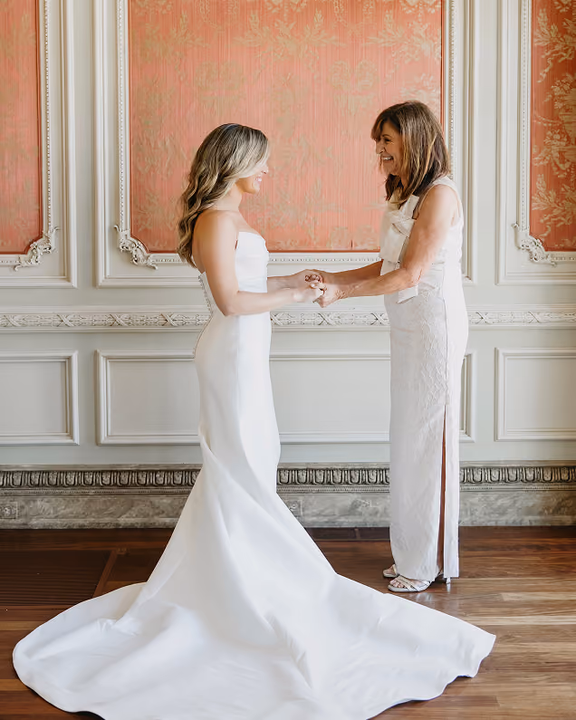 Bride in a strapless white wedding gown smiling and holding hands with a woman in a sleeveless white dress in an elegant room with ornate pink and white walls.