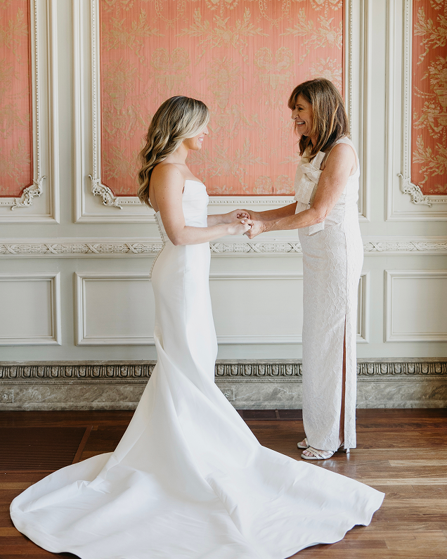 Bride in a strapless white wedding gown smiling and holding hands with a woman in a sleeveless white dress in an elegant room with ornate pink and white walls.
