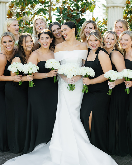 Bride in white off-shoulder gown holding white rose bouquet surrounded by bridesmaids in black dresses holding white rose bouquets, all smiling outdoors.