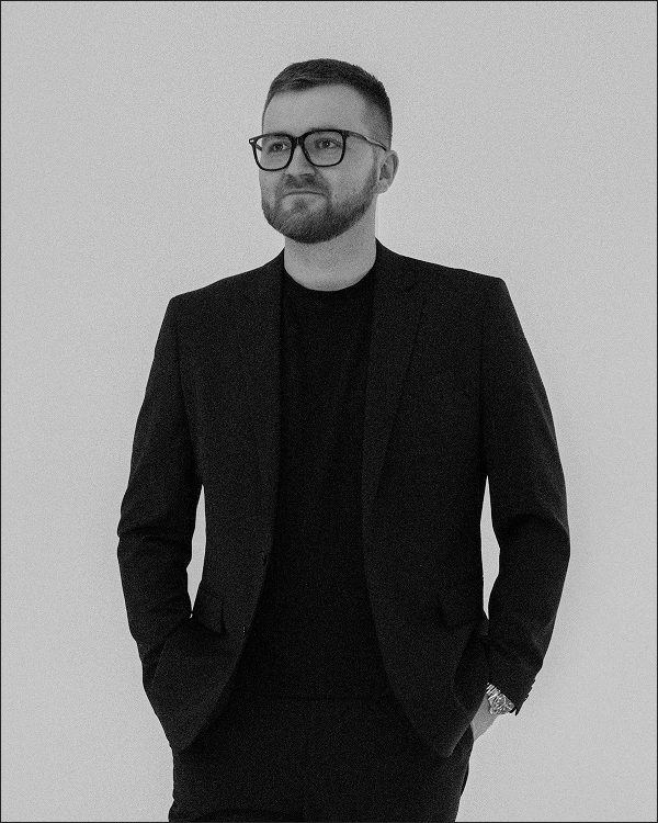 Man with short hair and beard wearing black glasses and a black suit with hands in pockets, looking to the side.