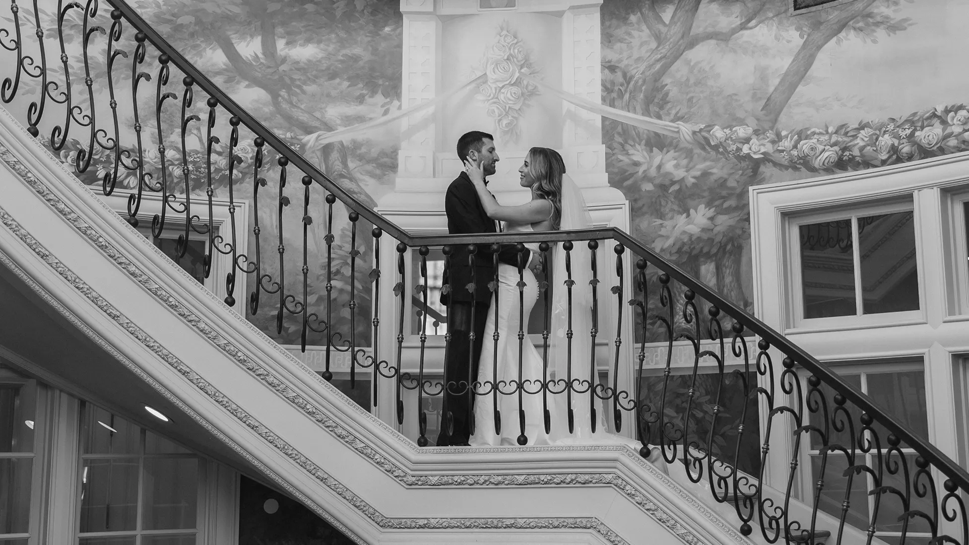 Bride and groom standing close on a decorative staircase with floral mural background, smiling at each other.