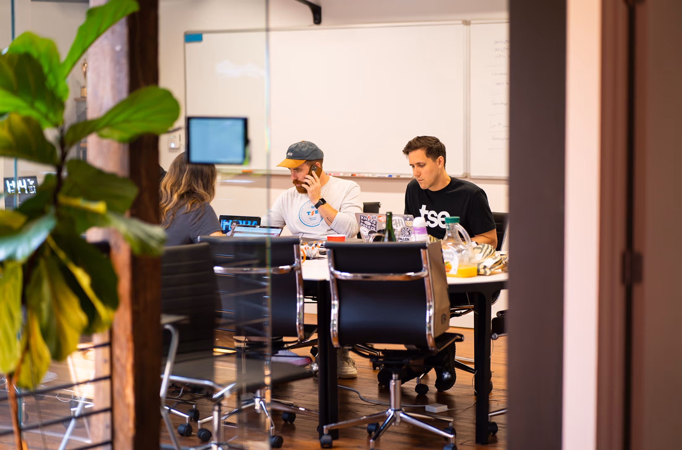 Three people working together at a conference table with laptops, one person on the phone, in a modern office with a large plant and whiteboard.