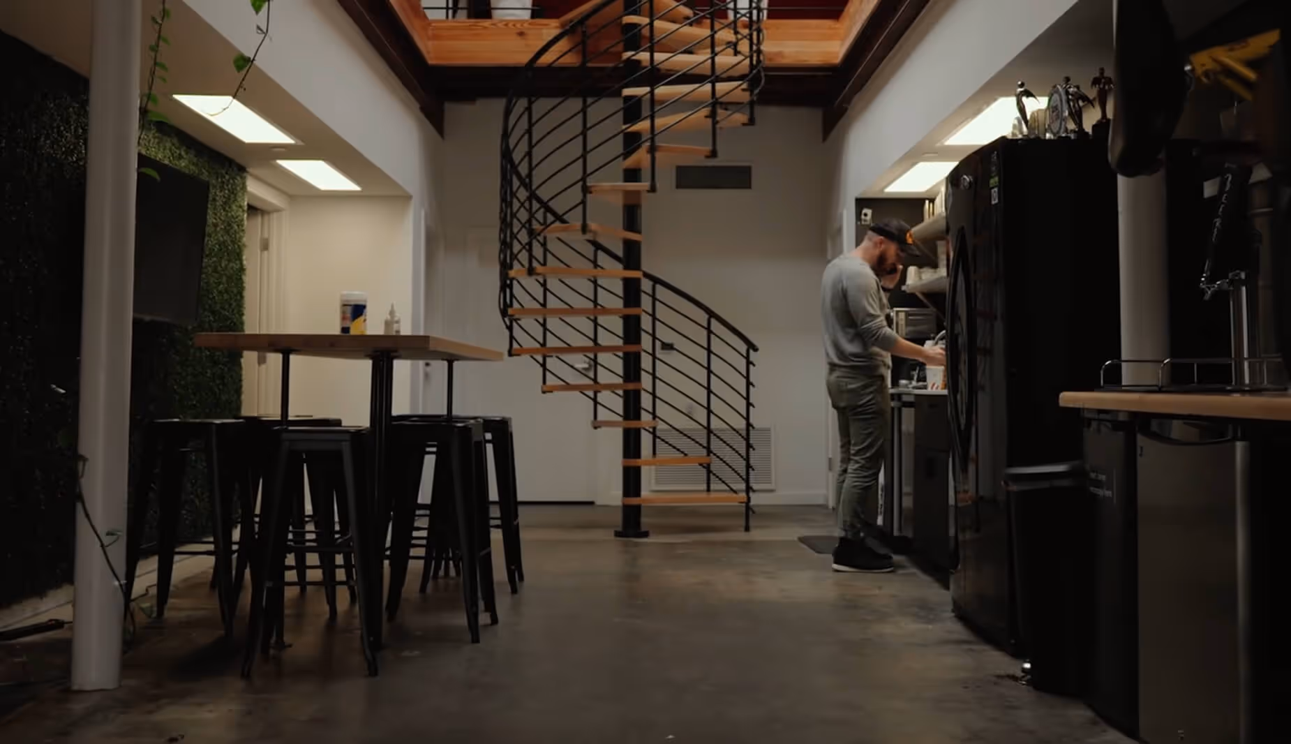Jeremy stands by a kitchen area in a modern office with a spiral staircase and high tables with stools.
