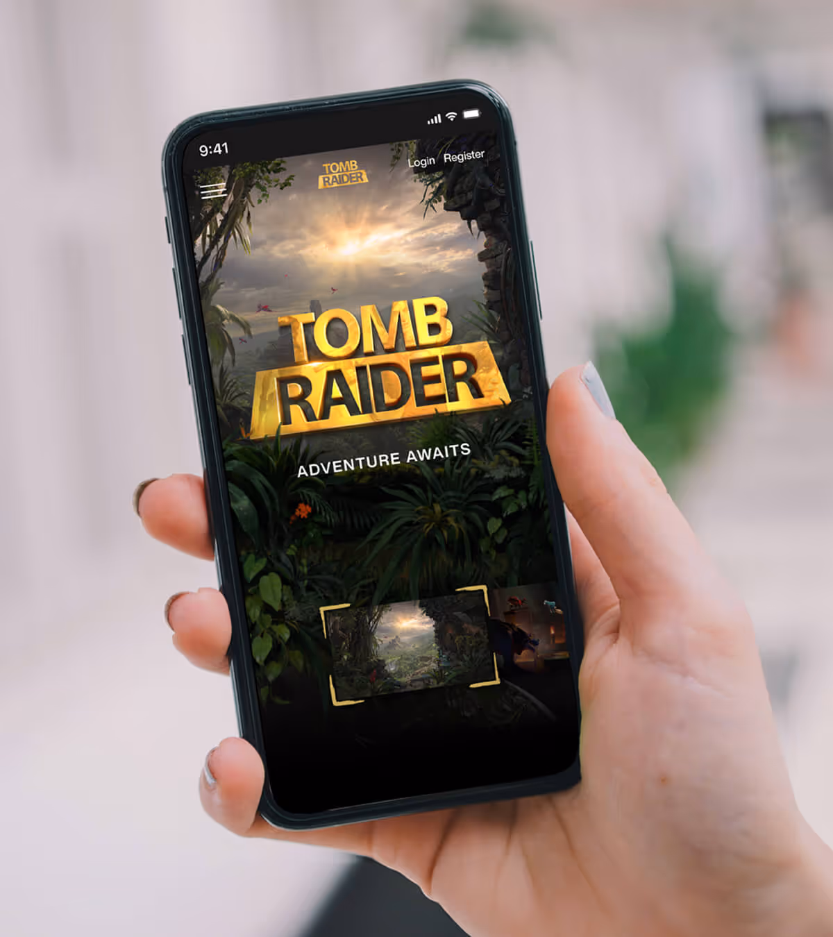 Hand holding a smartphone displaying the Tomb Raider app with 'Adventure Awaits' text on the screen.