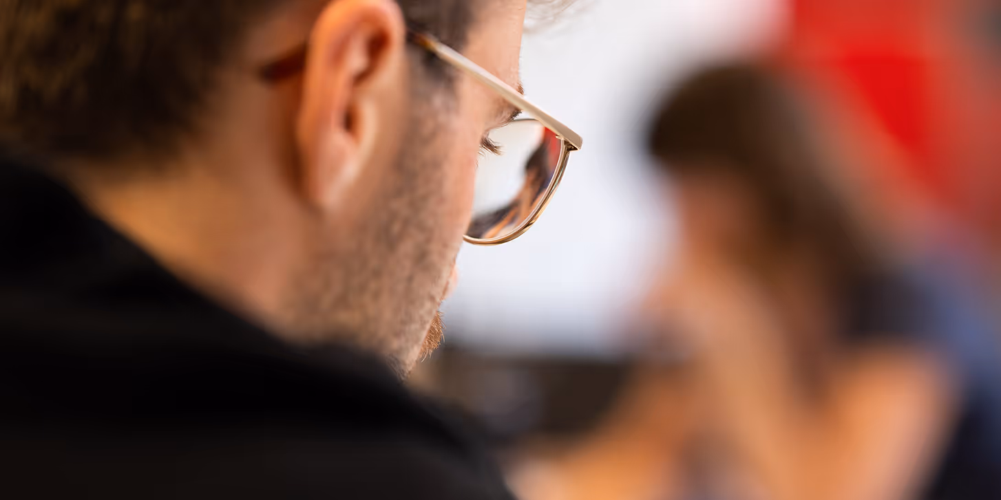 Close-up side view of a man with glasses and curly hair looking downward, with a blurred background showing a person in blue.