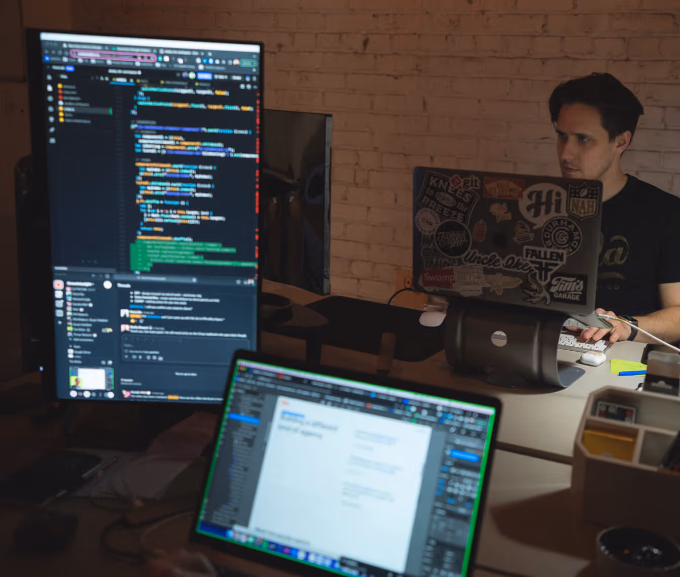Person working on a laptop covered with various stickers next to a large monitor displaying code and a blurred laptop screen in the foreground.