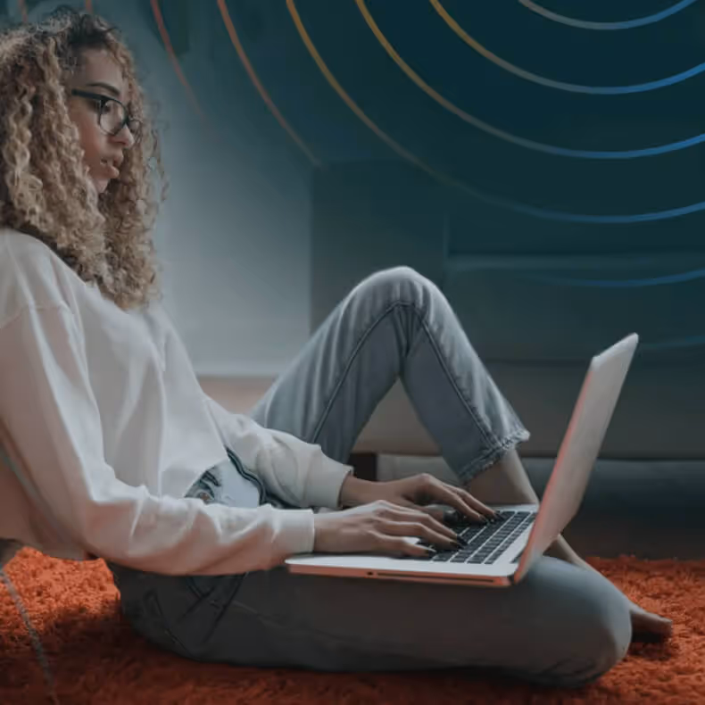 Woman with curly hair and glasses sitting on an orange carpet using a laptop.