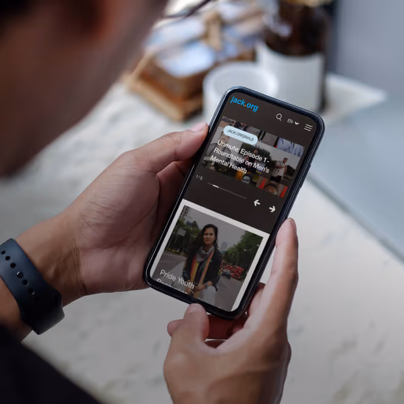 Person holding a smartphone displaying the jack.org website with a video titled 'Episode 1: Roundtable on Men's Mental Health' and another image titled 'Pride Youth'.