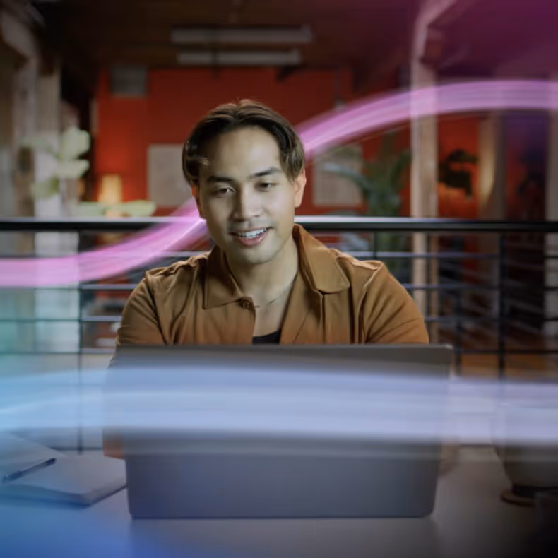 Young man with brown jacket working on a laptop in a modern office with colorful light trails.