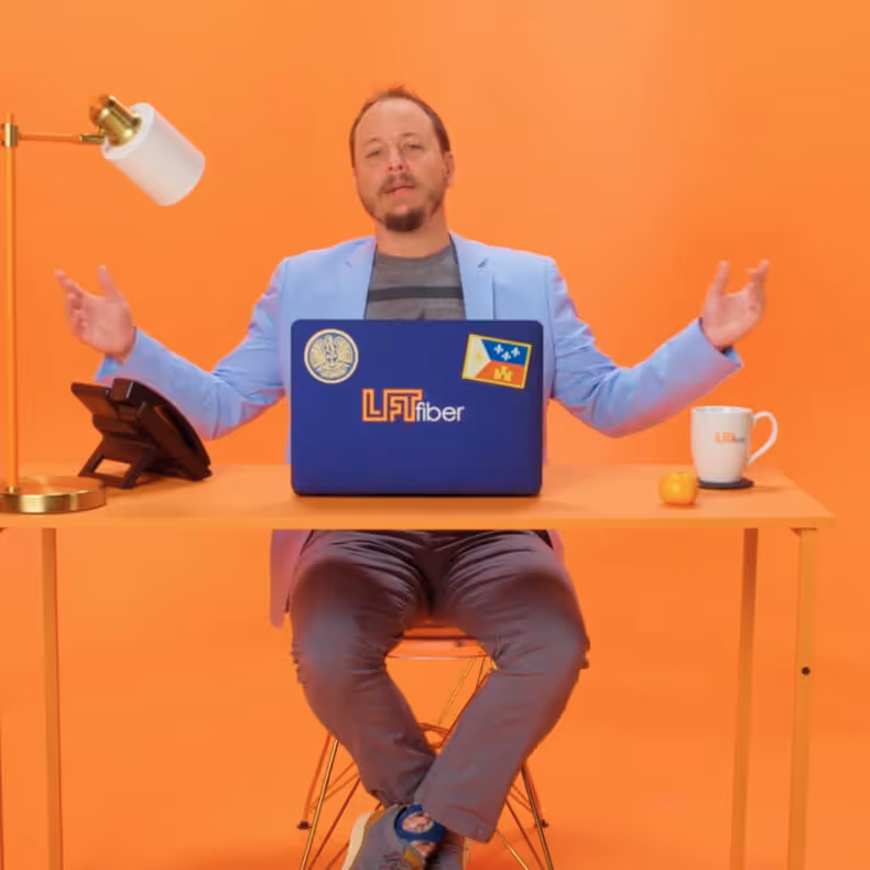 Man sitting at orange desk with laptop covered in LFT Fiber stickers, a lamp, phone, mug, and an orange against an orange background.
