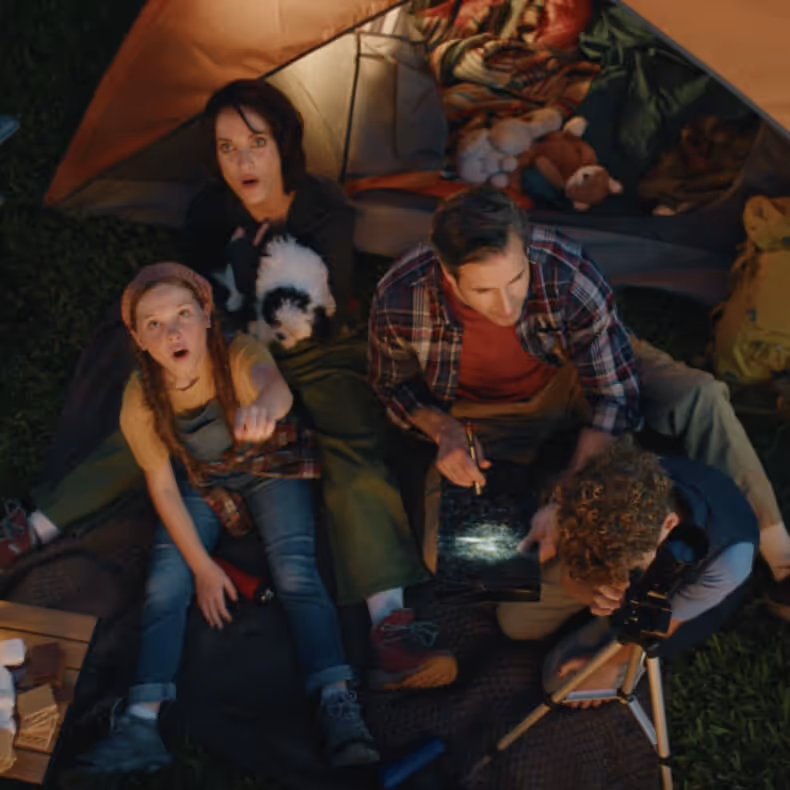 Four people sitting outside a tent at night, one pointing up, with a small black-and-white dog and a telescope nearby.