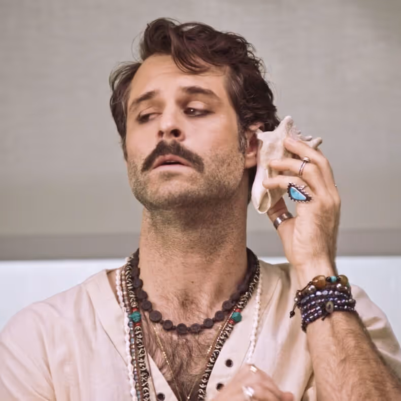Man with mustache holding a seashell to his ear, wearing multiple necklaces, rings, and bracelets.