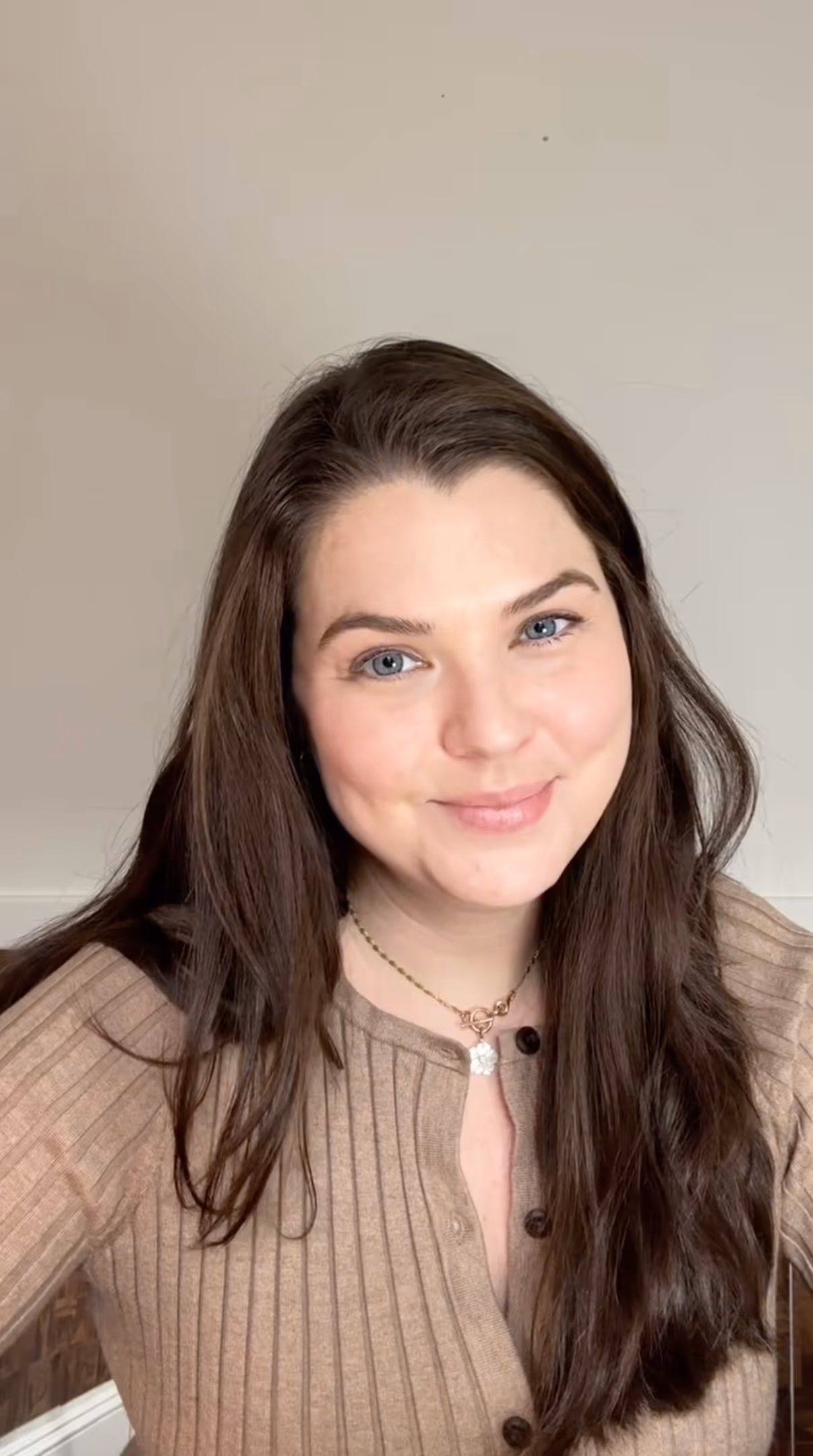 Smiling woman with long brown hair wearing a beige ribbed sweater and a gold necklace with a flower pendant.