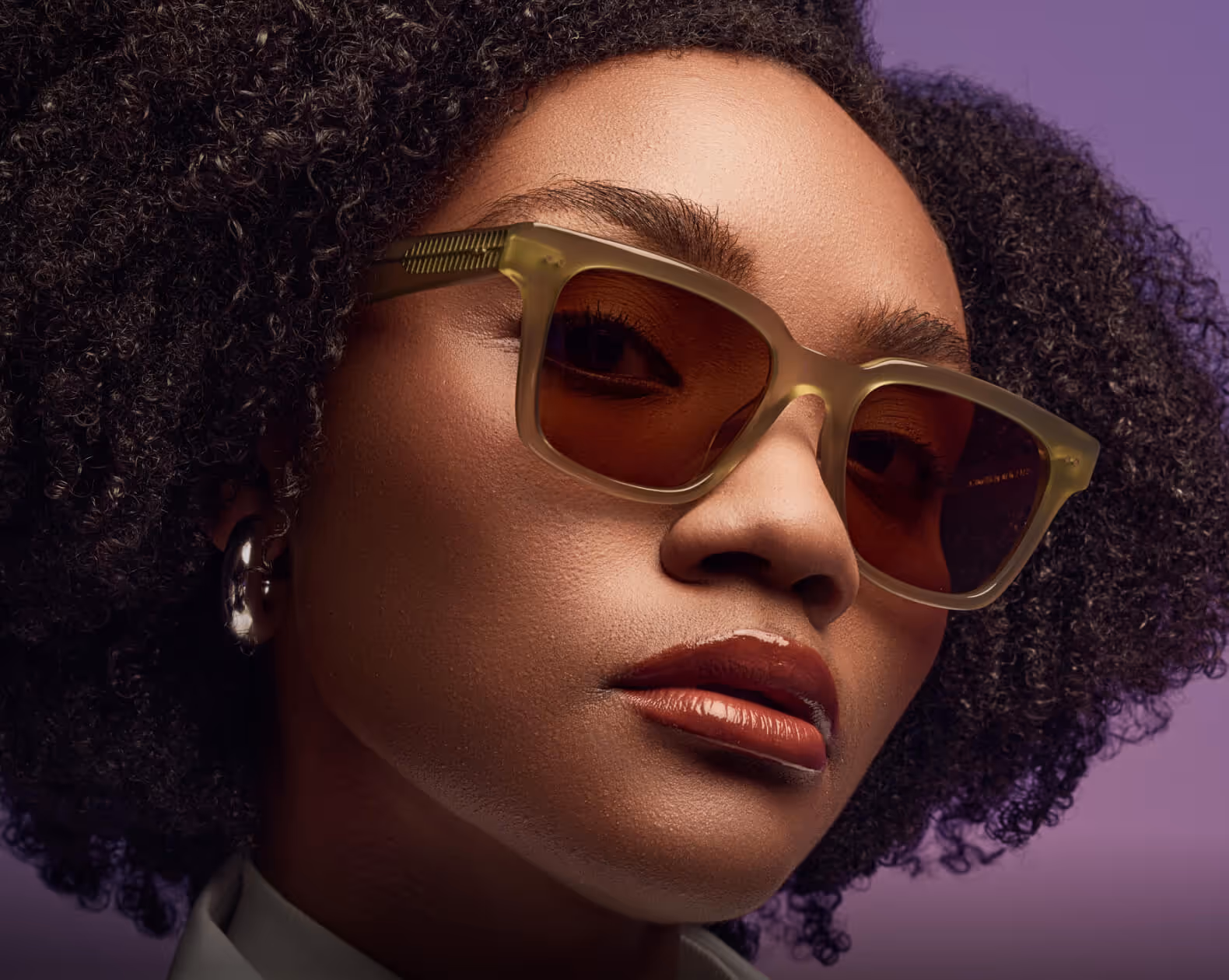 Close-up of a woman with curly hair wearing translucent green sunglasses with brown lenses and glossy dark red lipstick against a purple background.