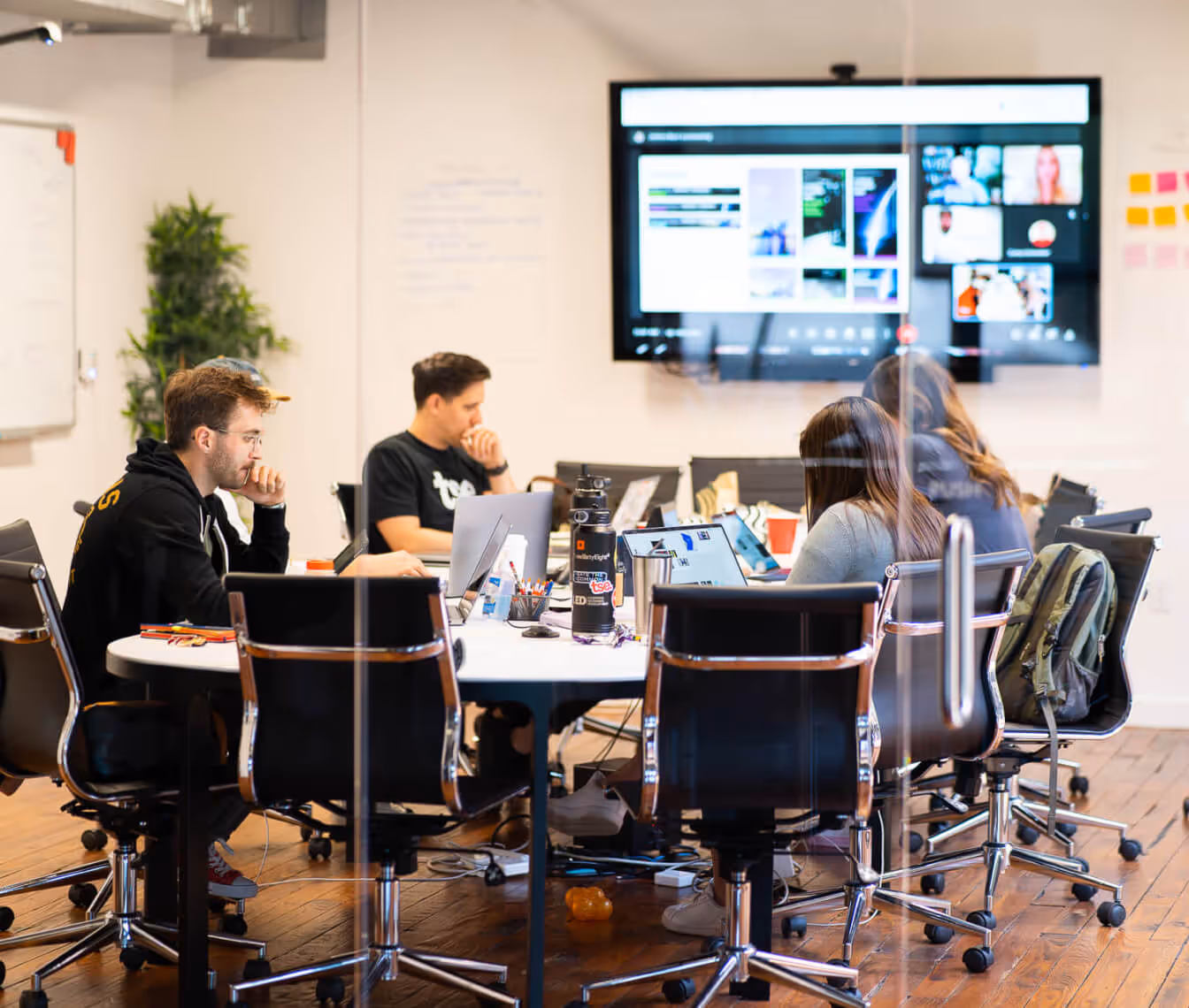Group of four people working on laptops around a conference table in a modern office with a large screen displaying a virtual meeting in the background.