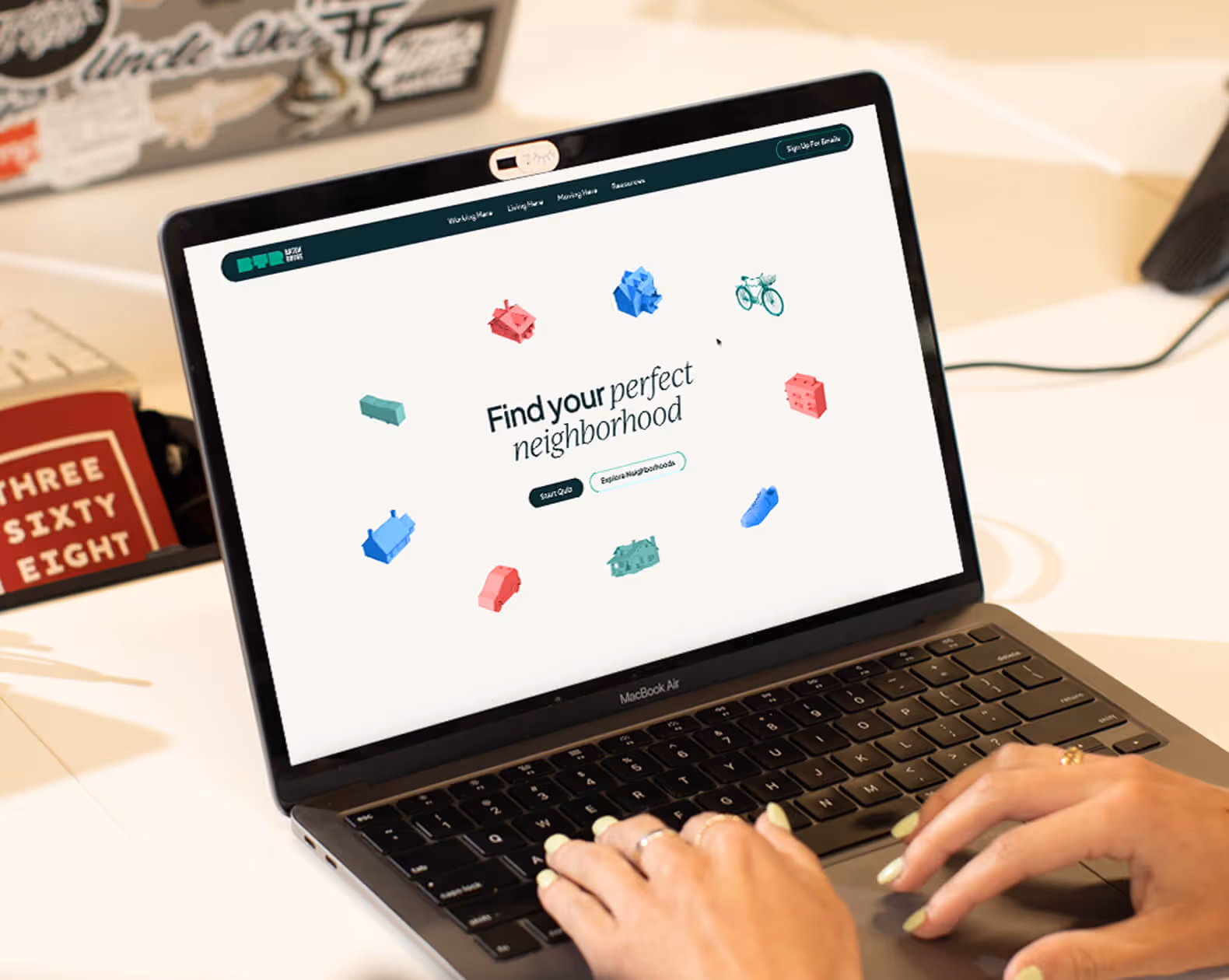 Person typing on a MacBook Air displaying a website with the text 'Find your perfect neighborhood' surrounded by colorful 3D icons of houses, a bike, a shoe, and other objects.