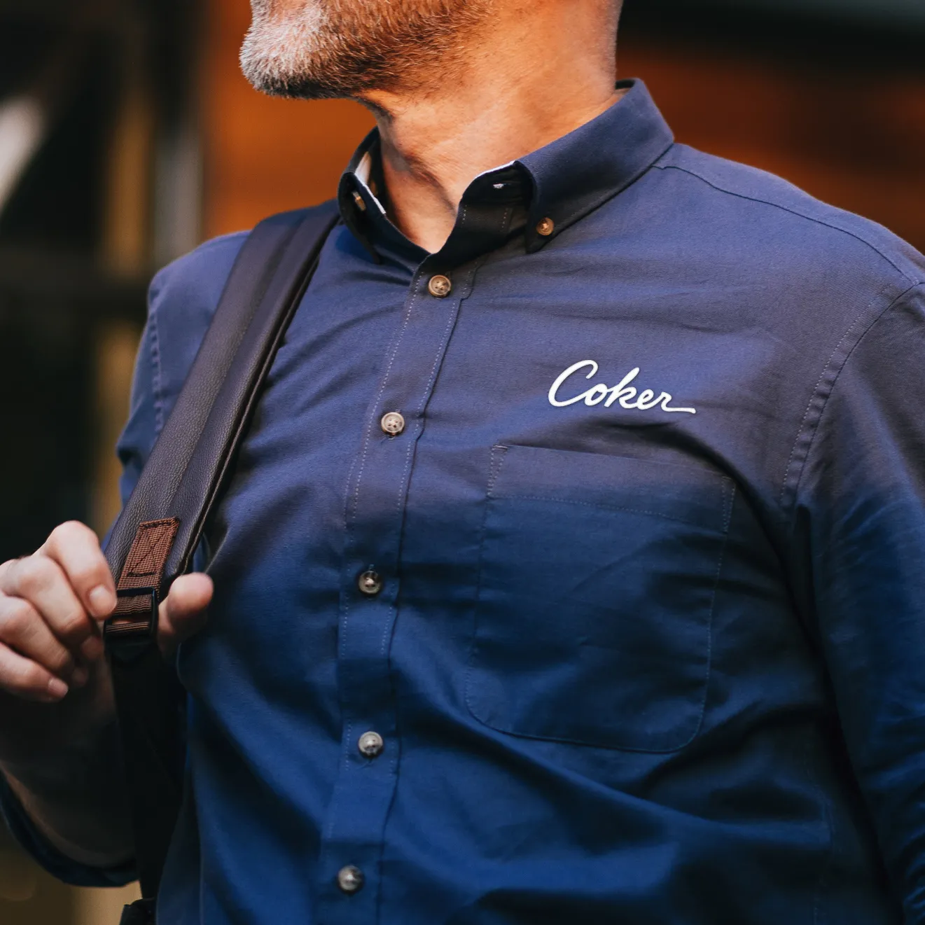 Close-up of a man wearing a blue button-up shirt with the word 'Coker' embroidered above the chest pocket and carrying a black backpack strap.